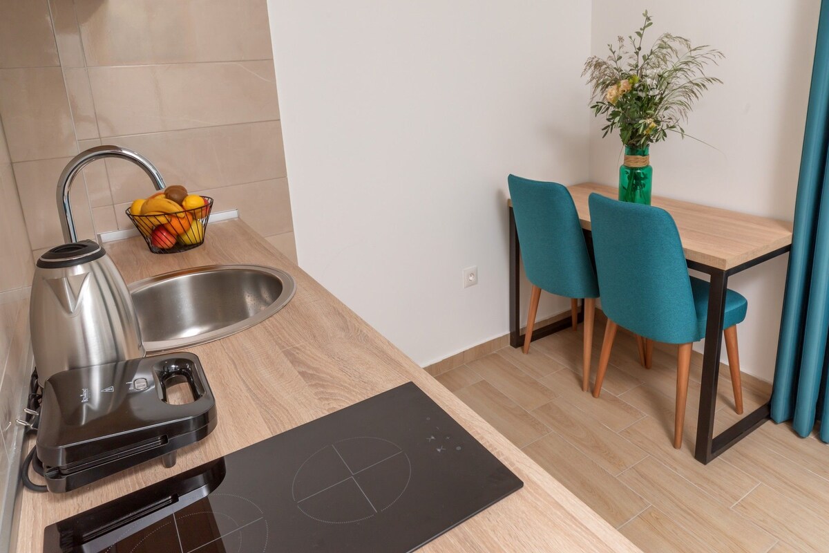 Cocina compacta con pileta, tetera y cocina por inducción. Hay una cesta de frutas en la mesada y se ve un comedor con dos sillas y una mesa.