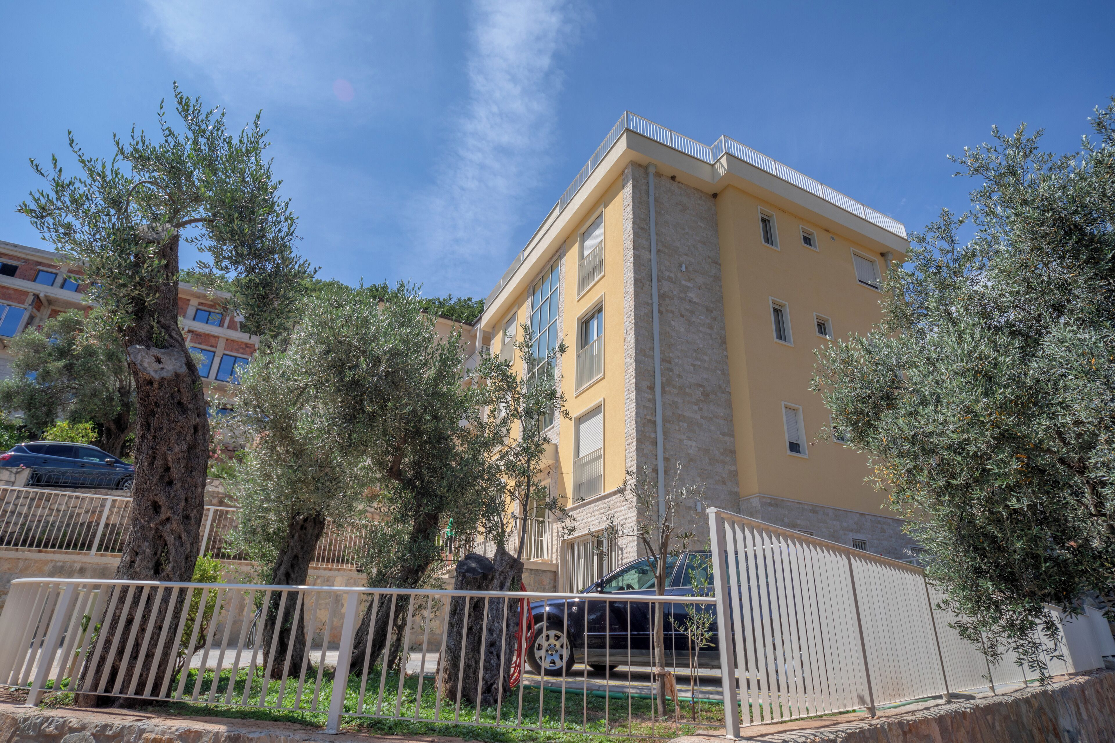 A yellow building with multiple windows and balconies, surrounded by trees and a white fence.