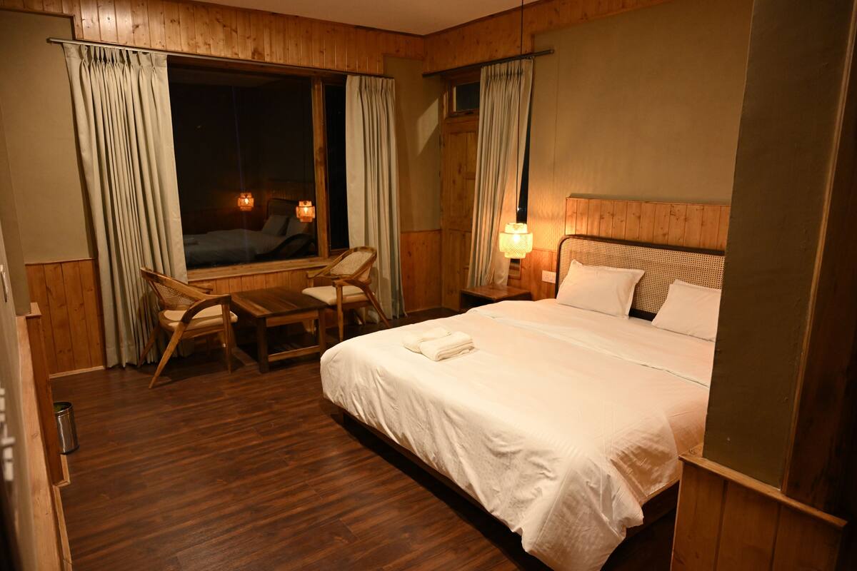 A hotel room with a large bed, a wooden headboard, a window with curtains, a desk with a chair, and two bedside lamps.