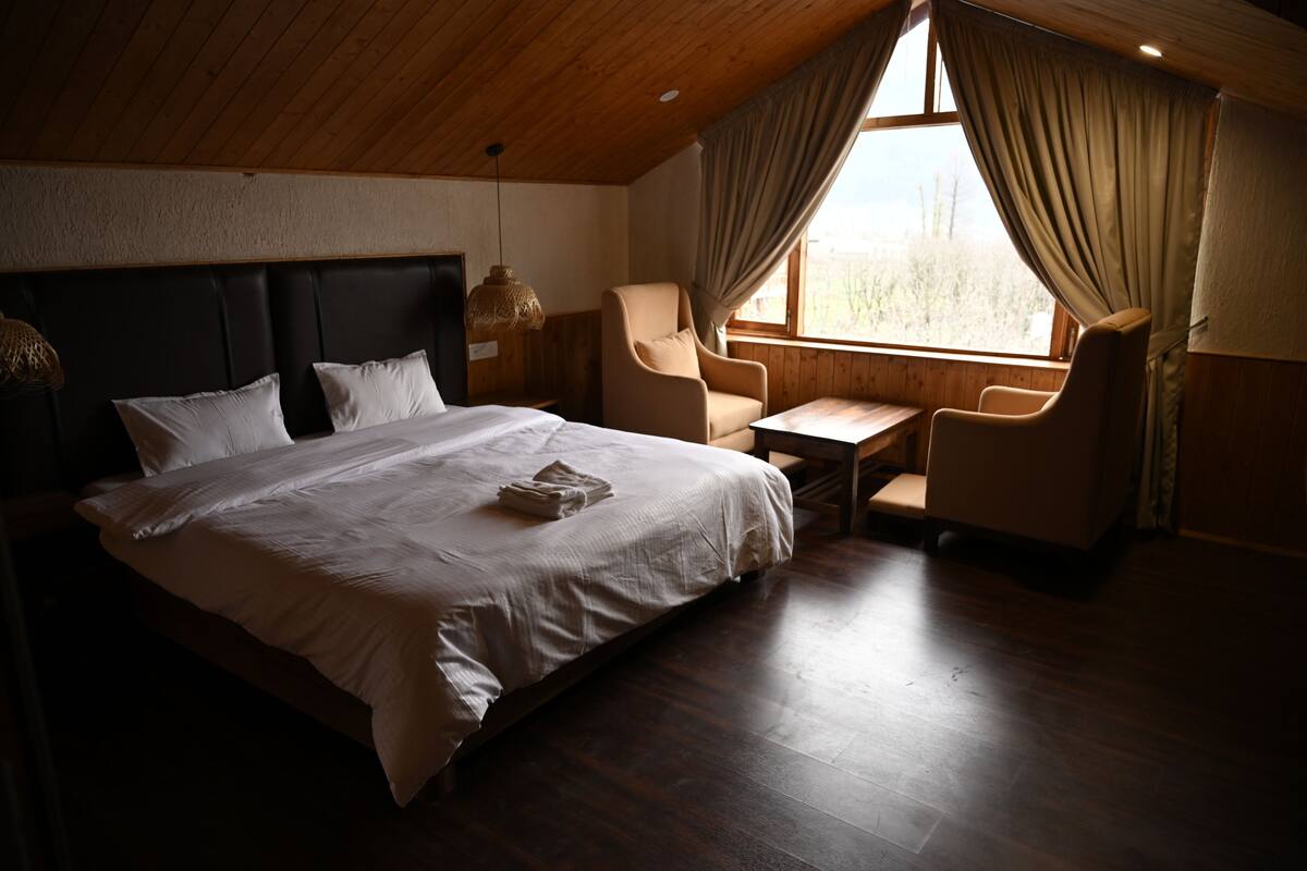 A bedroom with a large bed, a small desk, a chair, and a window with curtains.