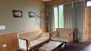 Una habitación con suelo de madera, un sofá beige, una mesa de centro de madera y una silla. Hay dos cuadros en la pared.