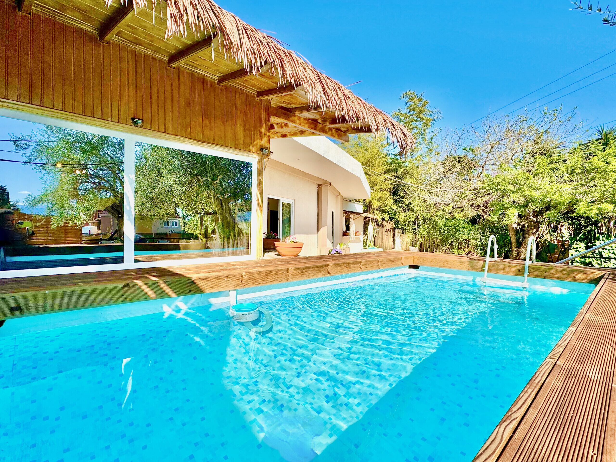 A swimming pool with a wooden deck, a thatched roof structure, and a view of a house in the background.