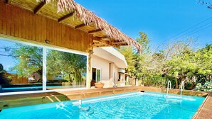 En swimmingpool med ett trädäck, en byggnad med halmtak och utsikt över ett hus i bakgrunden.