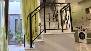 A modern kitchen with a built-in washing machine, a staircase with ornate black railings, and a large window with green curtains.