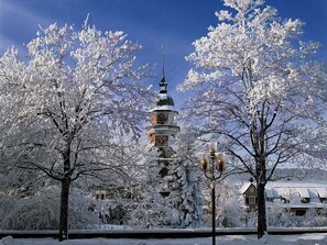 Ein mit Schnee bedeckter Turm mit einer Uhr, umgeben von Bäumen und einem Haus.