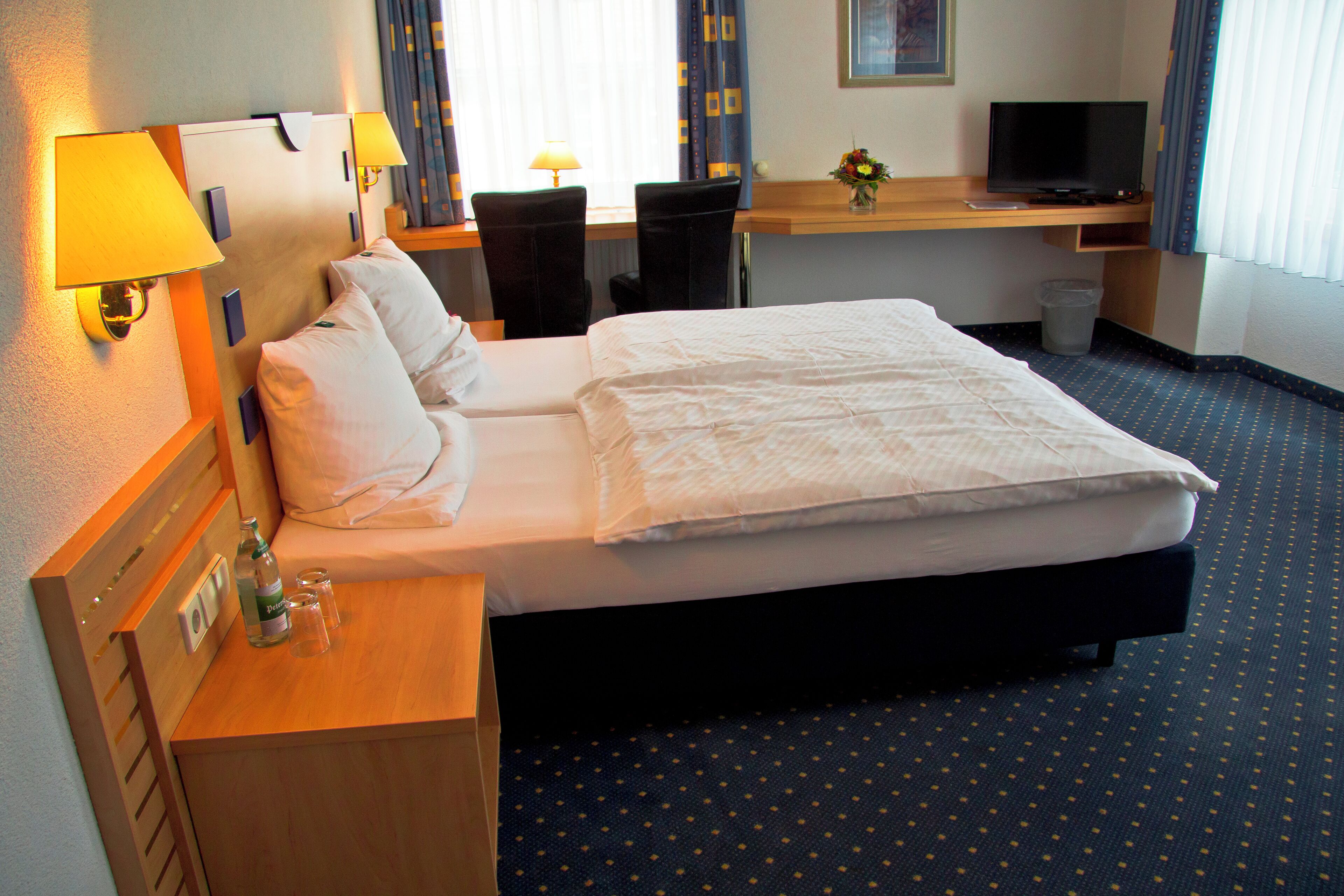 Una camera d'albergo con un letto, comodini, lampade, una scrivania con un computer e un cestino.