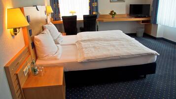 Una camera d'albergo con un letto, comodini, lampade, una scrivania con un computer e un cestino.