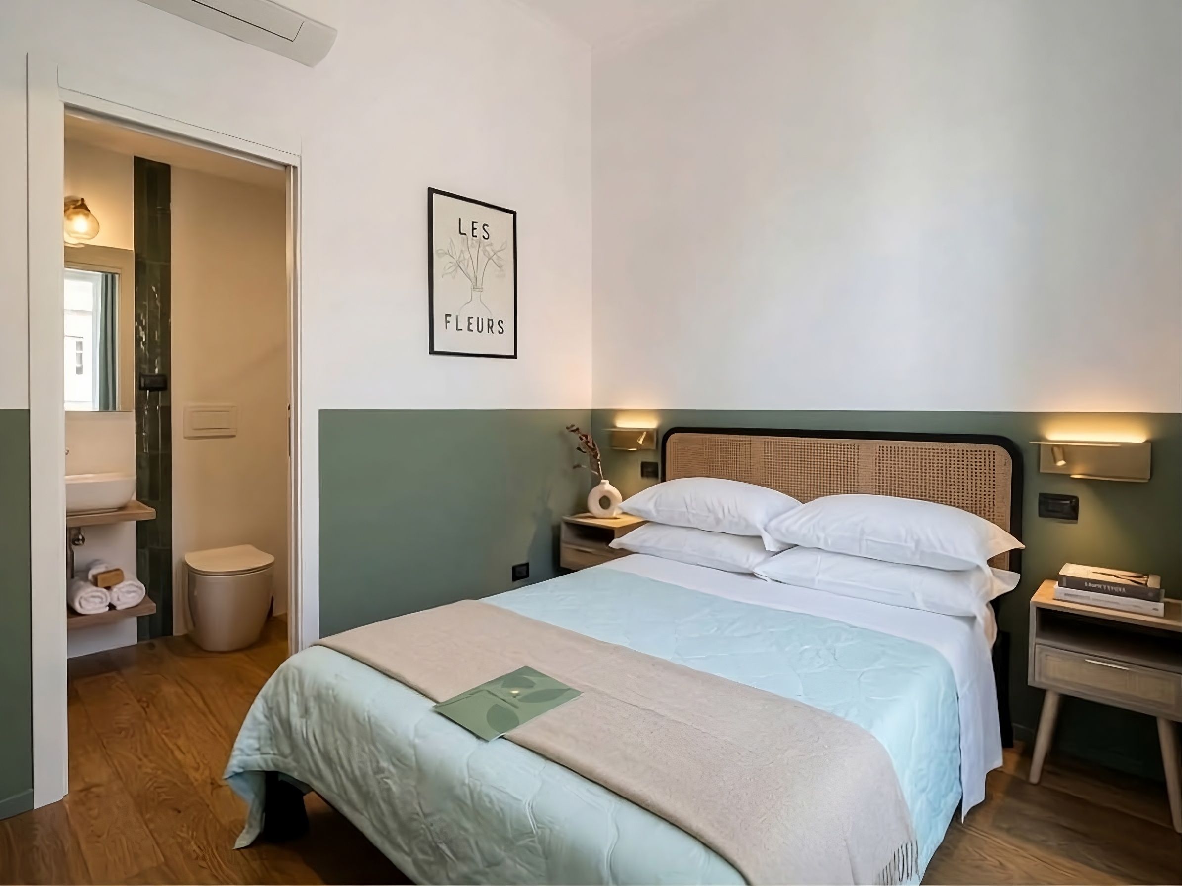 Ein Schlafzimmer mit einem Bett, einem Nachttisch, einem Schminktisch, einem gerahmten Bild an der Wand und einem durch eine offene Tür einsehbaren Badezimmer.