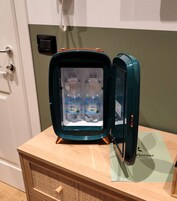 Een compacte, turquoise koelkast met glazen deur, waarin twee waterflessen staan met de aanduiding 'Soguaro'. De koelkast staat op een houten tafel tegen een groene muur, met een deur en een telefoon op de achtergrond.