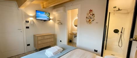Habitación de hotel con cama, televisor montado en la pared, cómoda de madera, baño con ducha y un adorno en la pared.