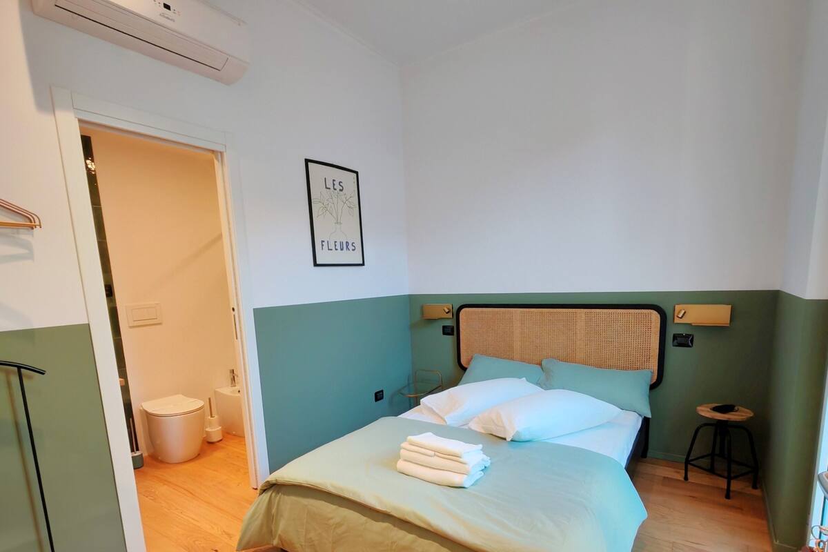 Een netjes opgemaakt slaapkamer met een bed, een klein krukje en een ingelijst beeld aan de muur.