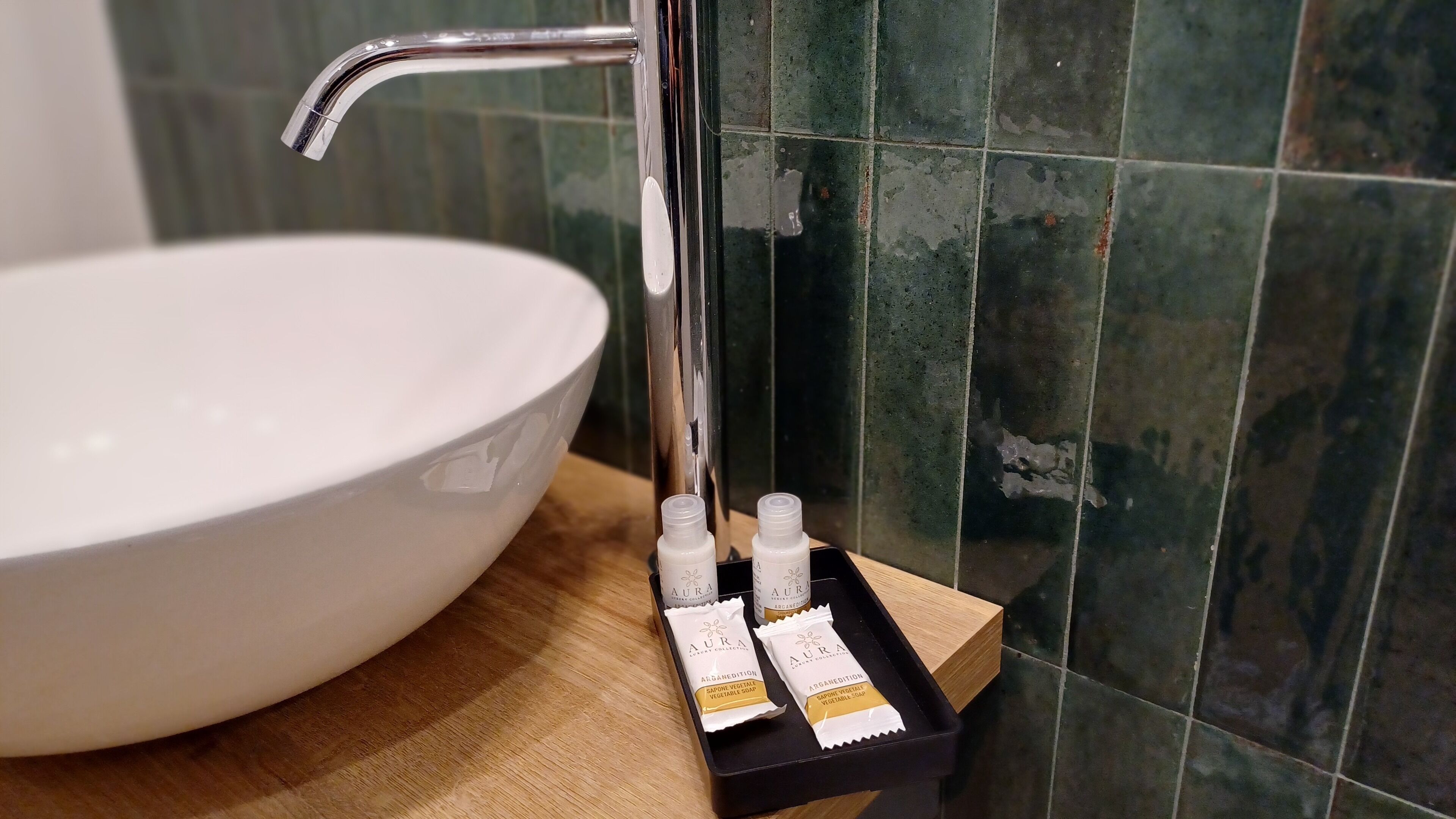 Ein Badezimmerwaschbecken mit einer weißen Schüssel und einem chromfarbenen Wasserhahn. Auf einer Holzschale stehen zwei Seifenflaschen mit der Aufschrift 'AURA'.