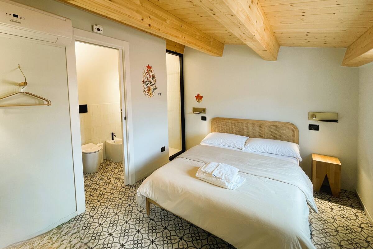 Een slaapkamer met een bed, houten plafond, een badkamer met toilet en wastafel, en een vloer met een patroon.