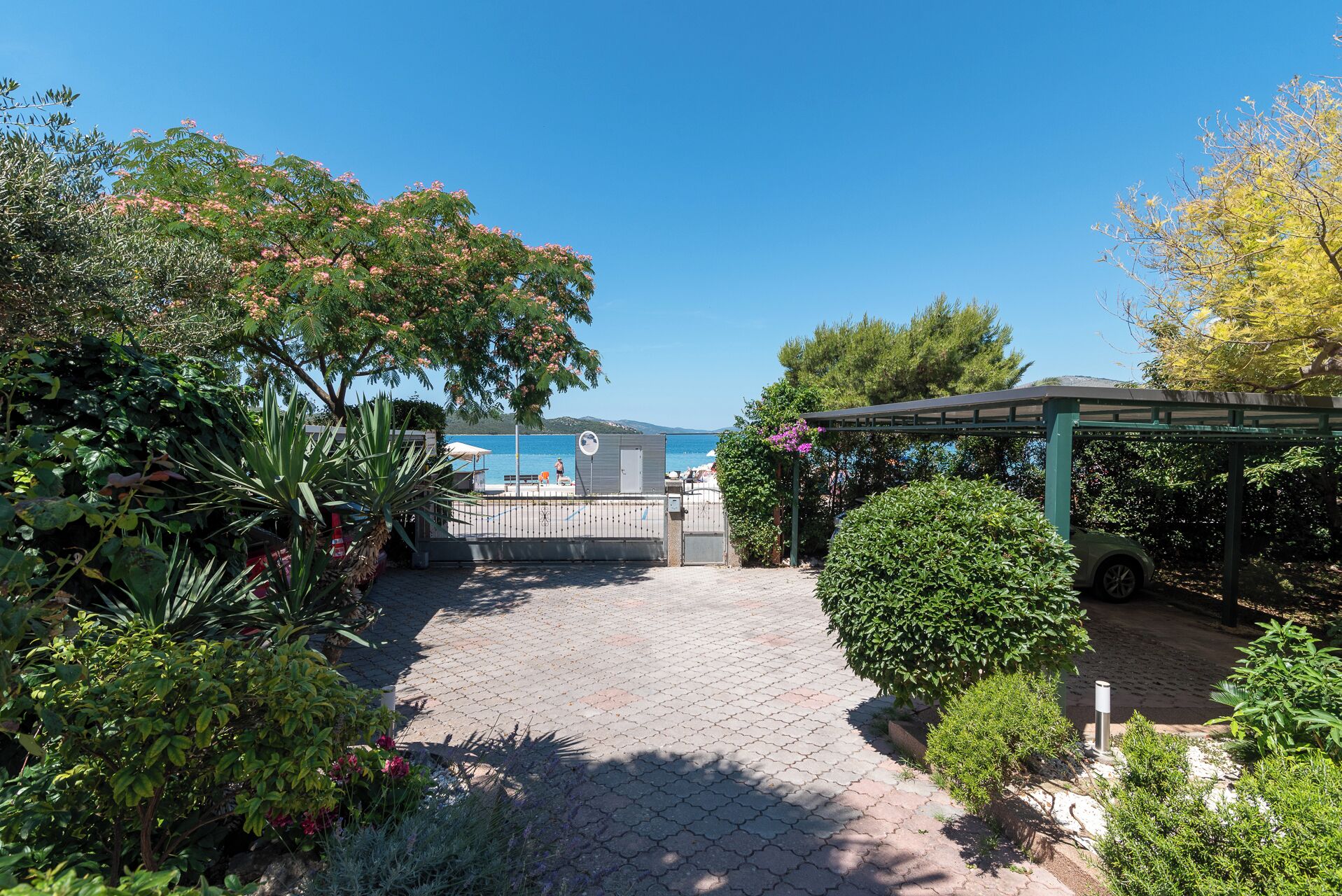 Un chemin pavé mène à une zone aménagée près de la plage, avec une pergola et une voiture garée à proximité.