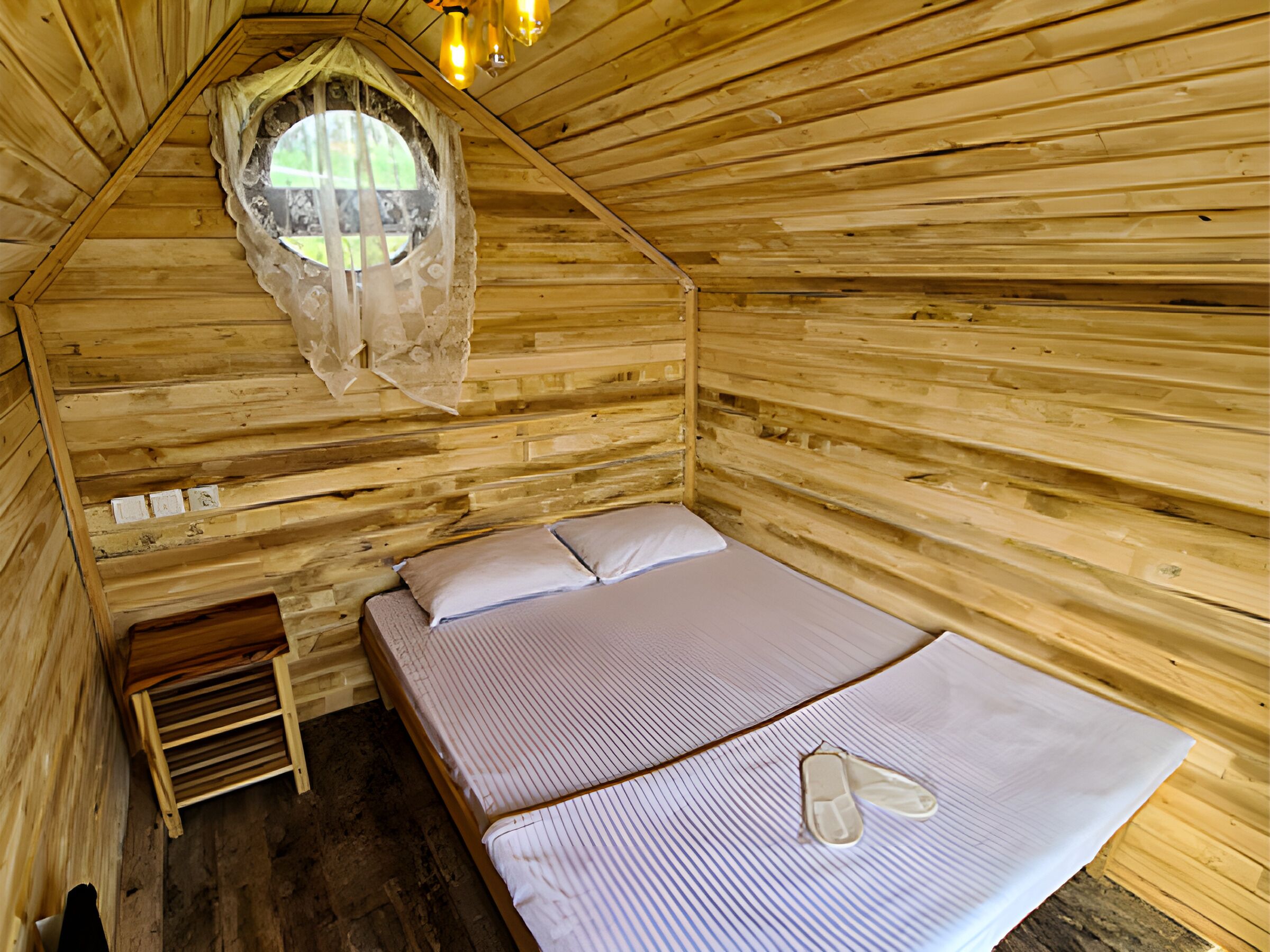 Habitación de cabaña de madera con cama, un pequeño banco de madera y una ventana con cortina de encaje.