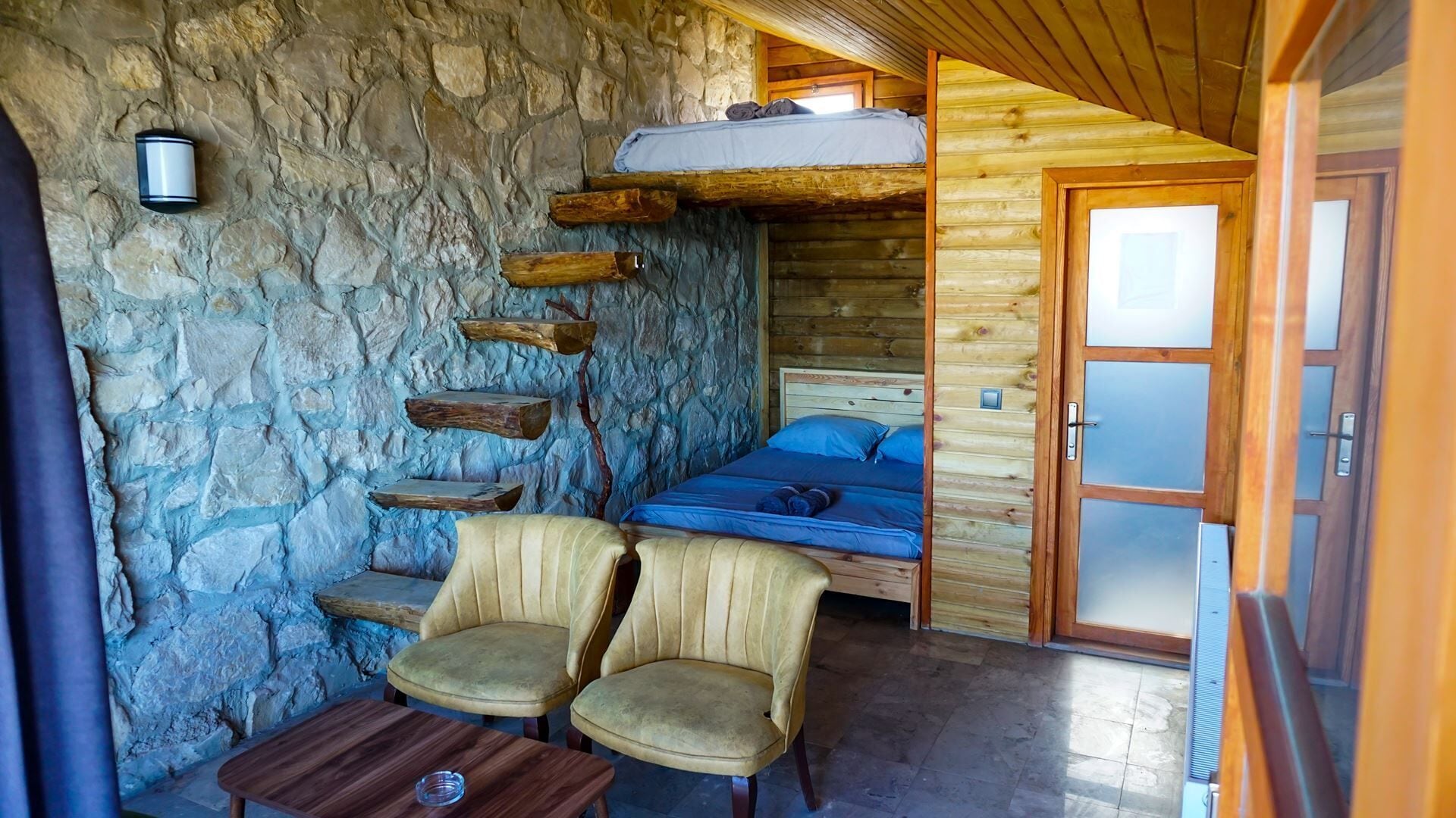 A rustic cabin interior with a stone wall, a bunk bed, two upholstered chairs, and a small wooden table.