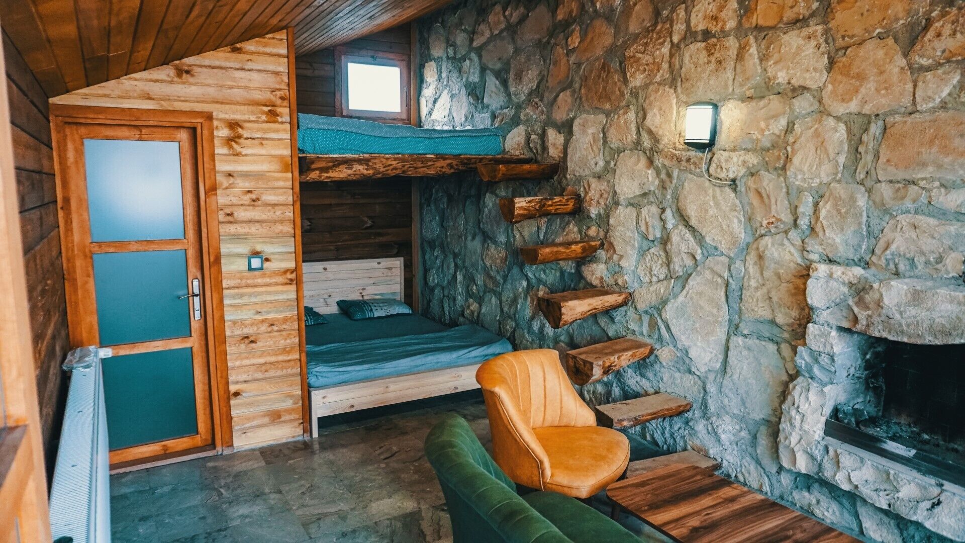 A cozy cabin interior with a stone wall, a wooden door, a bed, a chair, and a fireplace.