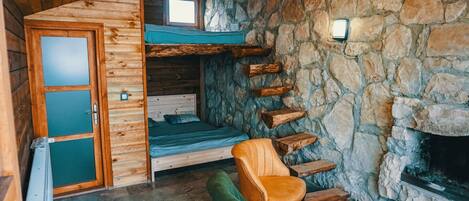 A cozy cabin interior with a stone wall, a wooden door, a bed, a chair, and a fireplace.