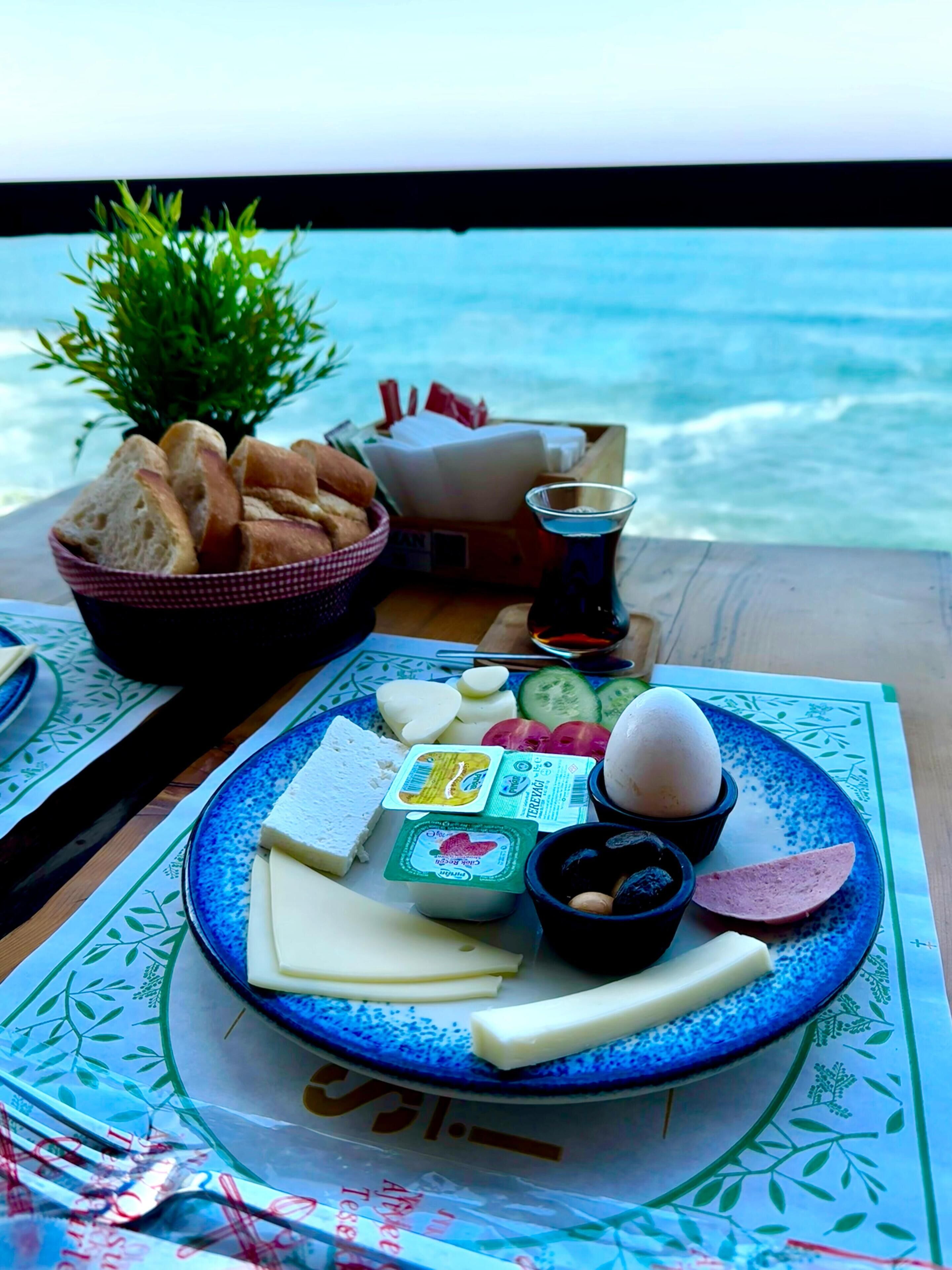 A table with a plate of cheese, eggs, and bread, a small plant, and a glass of water, set against a view of the sea.