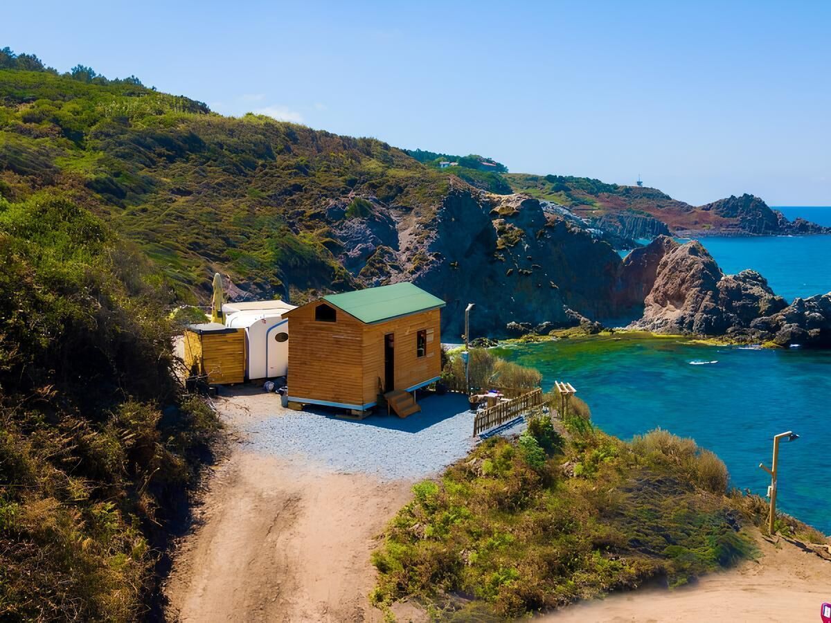 Una piccola cabina di legno con tetto verde, una toilette chimica e un vialetto sterrato che conduce all'acqua.