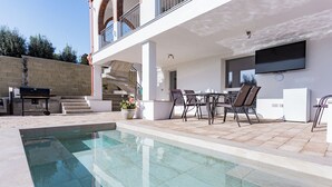 A modern outdoor pool area with a barbecue, seating, and a TV mounted on the wall. - Villa with heated pool 2 steps from the sea (Quartu Sant'Elena)