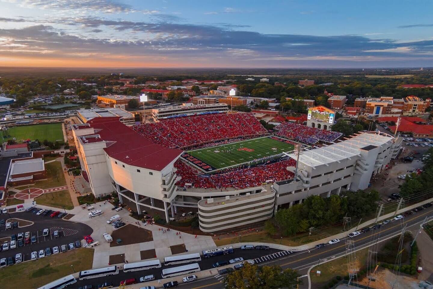 An aerial view of a stadium with a football field, surrounded by buildings and parking areas.