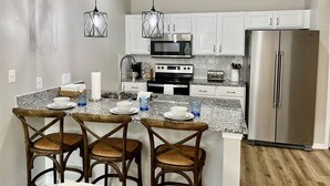 Cocina moderna con armarios blancos, electrodomésticos de acero inoxidable y encimera de granito. Hay tres sillas de madera con asientos trenzados y una pequeña isla con un lavamanos y algunos objetos sobre la encimera.