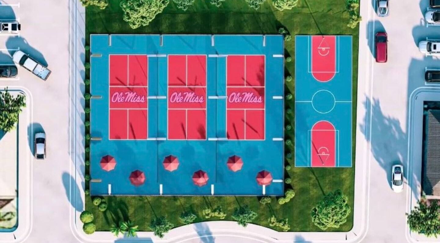 An aerial view of a sports complex with multiple basketball courts, parking areas, and surrounding greenery.