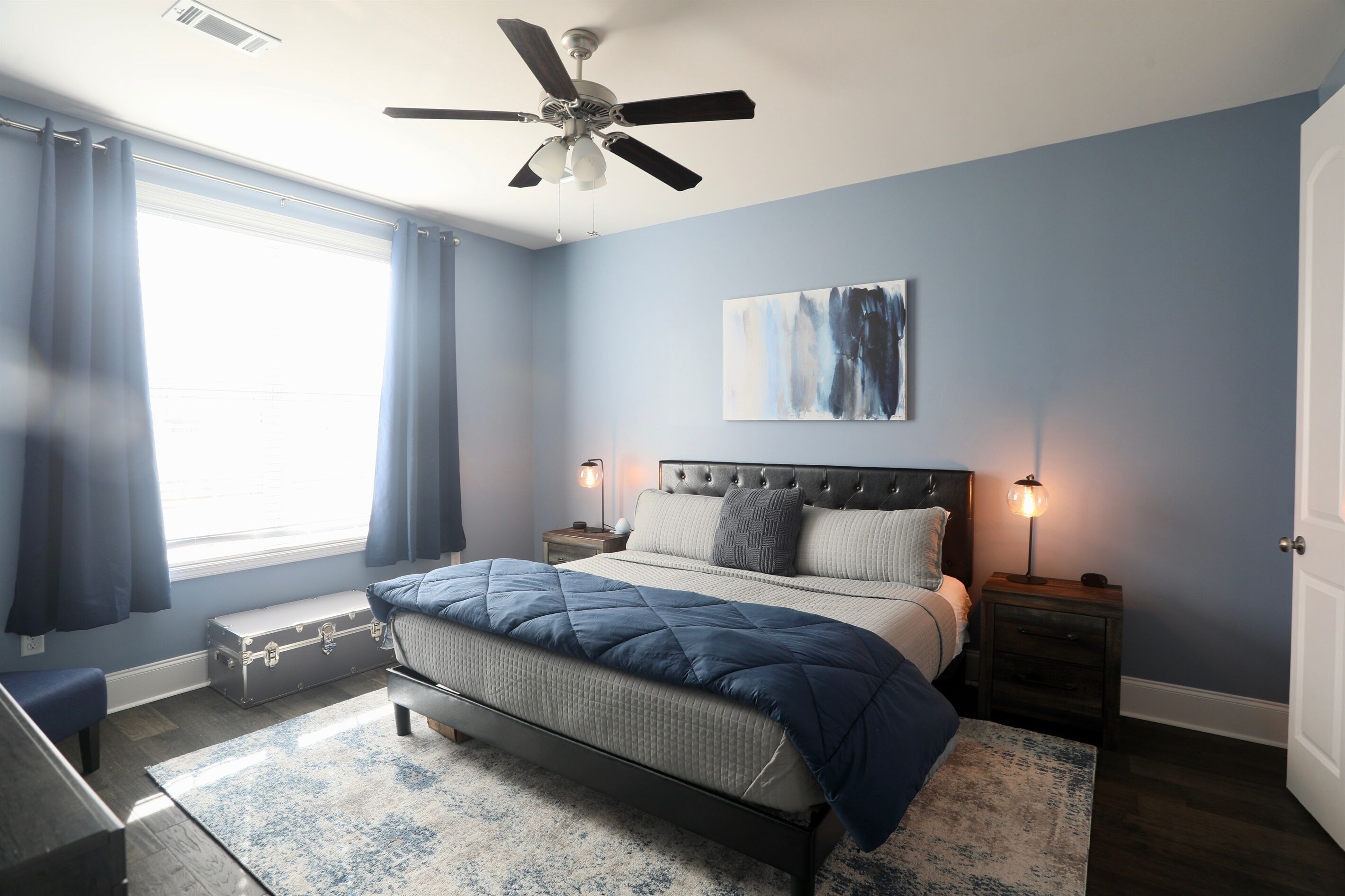 A bedroom with a bed, a ceiling fan, a nightstand, a bedside lamp, and a window with curtains.