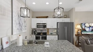 Una cocina moderna con encimeras de granito, muebles blancos, un refrigerador de acero inoxidable, microondas y estufa a gas. Hay dos lámparas colgantes, un lavaplatos y un florero sobre la encimera.