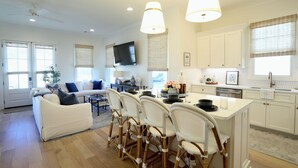 A modern kitchen with white cabinetry, a dining area with wicker chairs, and a living room with a white sofa.