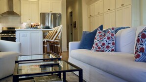 A modern living room with a white sofa, a glass coffee table, and a kitchen in the background.