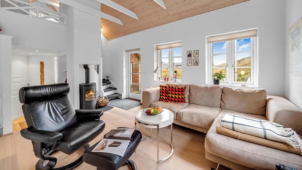 A modern living room with a black leather sofa, a beige sofa, a round coffee table, and a black leather chair. There is a fireplace, a window with a view, and a door leading to another room. - Modernized holiday home, centrally located in one of Søndervig's best locations (Ringkøbing)