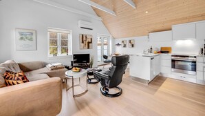 A modern living room with a sofa, a black chair, a dining table, and a kitchen area with white cabinets and appliances. - Modernized holiday home, centrally located in one of Søndervig's best locations (Ringkøbing)