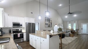 Une cuisine spacieuse dotée de meubles blancs, d’appareils électroménagers en acier inoxydable, de plans de travail en granit et de luminaires suspendus.