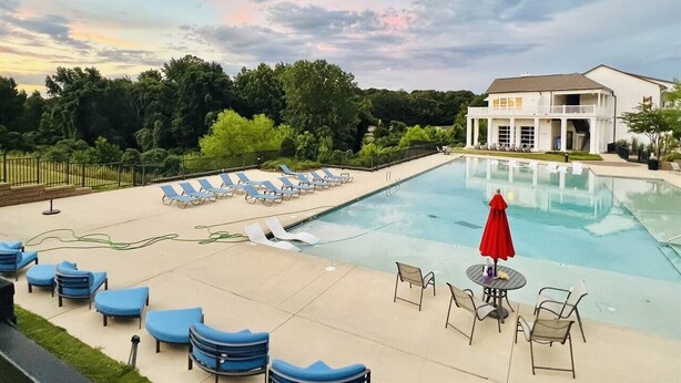 Une piscine entourée de chaises longues et de parasols, le tout bordé de verdure, avec un bâtiment visible au loin.