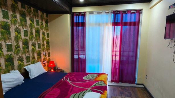 A bedroom with a bed, a nightstand, a wall-mounted TV, and a window with curtains. - Hotel Pangot Himalayan View (Kosiyakutoli)
