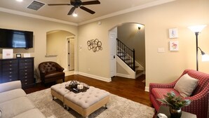 A living room with a ceiling fan, a flat-screen TV, a brown leather armchair, a red patterned armchair, a beige ottoman, and a white rug. - The Great Southern - Chic Oxford Getaway (Oxford)