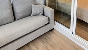 A modern living room with a grey sofa, a small table, and a sliding glass door leading to a bedroom with a bed.
