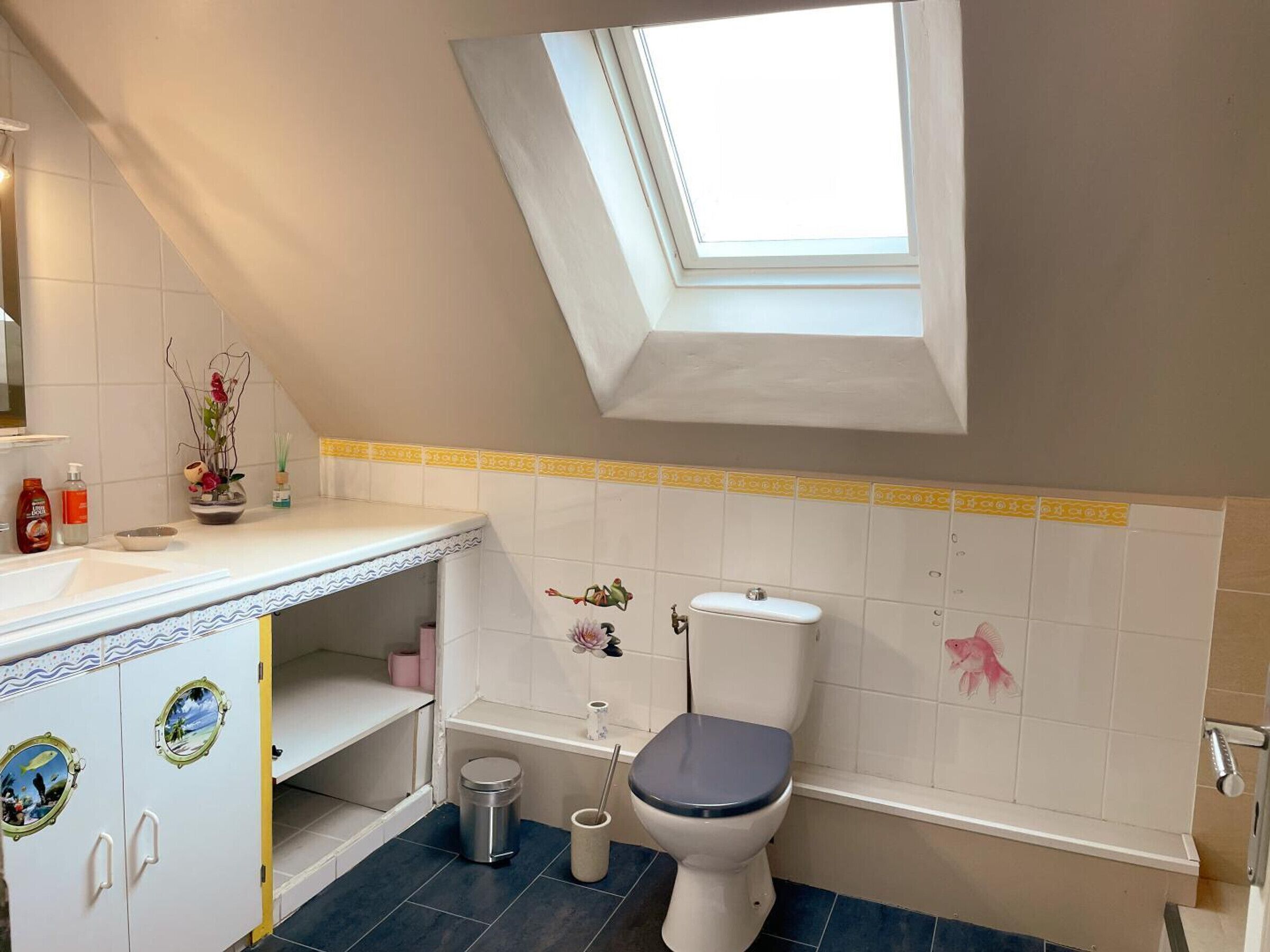 Een badkamer met dakraam, witte tegels, een toilet, een prullenbak en een wastafel met decoratieve objecten.