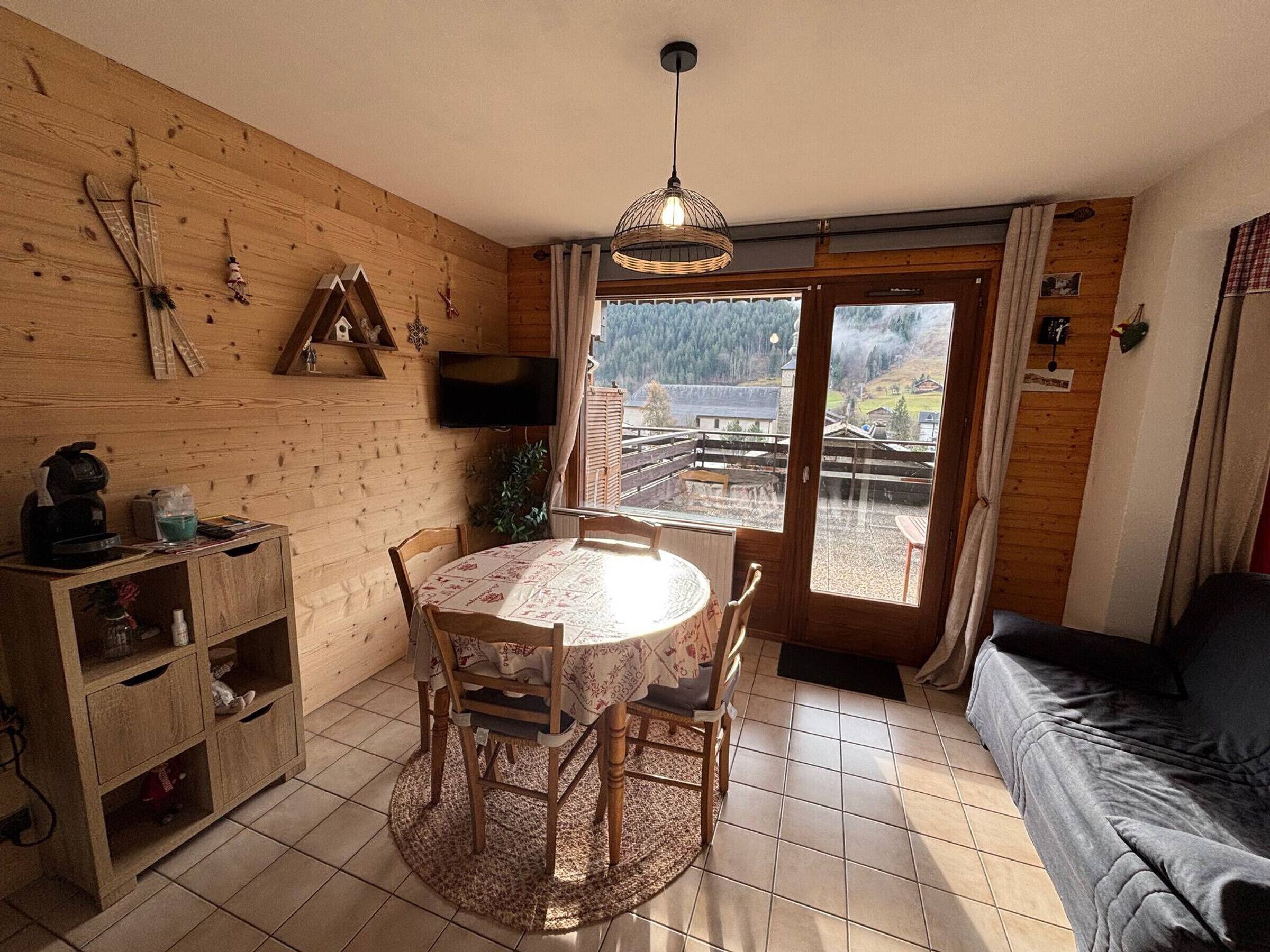 A cozy room with a wooden interior, a round dining table with chairs, a sofa, a TV mounted on the wall, and a view of a mountainous landscape through a glass door.