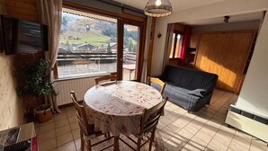 Un coin repas comprenant une table ronde, des chaises, un téléviseur et une vue sur un paysage rural grâce à de grandes fenêtres.