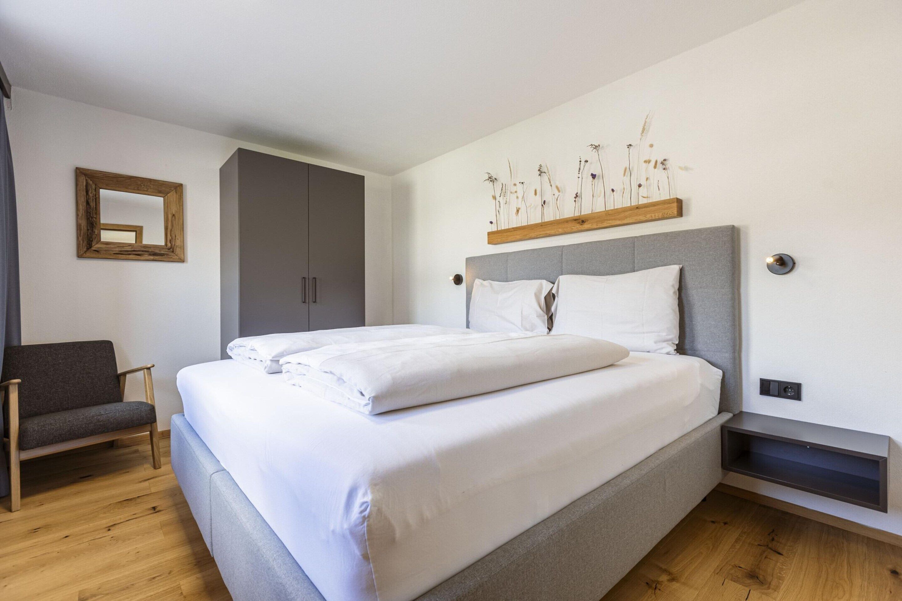 Een net opgemaakt bed met witte lakens, een grijs gestoffeerd hoofdbord en een houten plank erboven.