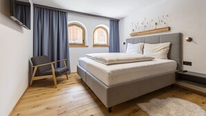 Une chambre moderne avec un lit, une chaise, deux fenêtres et un téléviseur fixé au mur.