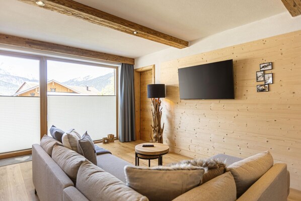A modern living room with a sofa, a round coffee table, and a flat-screen TV mounted on a wooden wall. - Apartment in Tyrol on the Ski-slope (Lermoos)