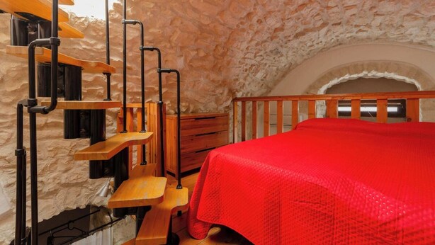 A room with a stone wall, a wooden bed with a red blanket, a wooden desk, and a staircase with black railings. - Little House in the Junno (Mattinata)