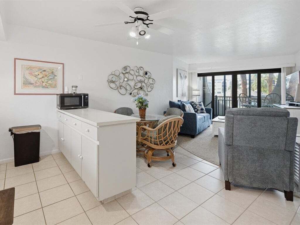 A modern kitchen with white cabinets, a microwave, and a dining area with a wicker chair and a grey sofa.