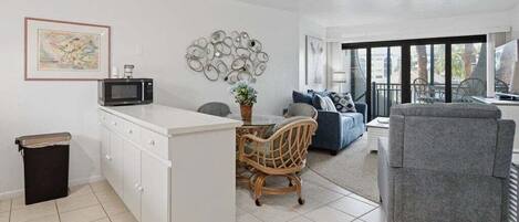 A modern kitchen with white cabinets, a microwave, and a dining area with a wicker chair and a grey sofa.