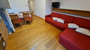 Un espace de vie compact comprenant un canapé rouge, une table à manger en bois, des chaises, un téléviseur fixé au mur et un plancher en bois.