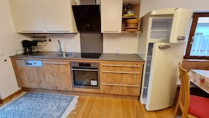 Une cuisine compacte équipée d’un lave-vaisselle, d’un four et d’une plaque de cuisson intégrée. On y trouve un petit évier et une bouilloire posés sur une étagère.